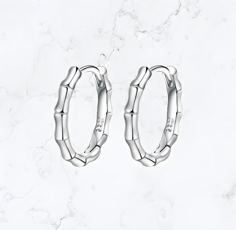 A pair of Gemineye Jewellery Silver Bamboo Huggies Earrings rest on a marble surface.